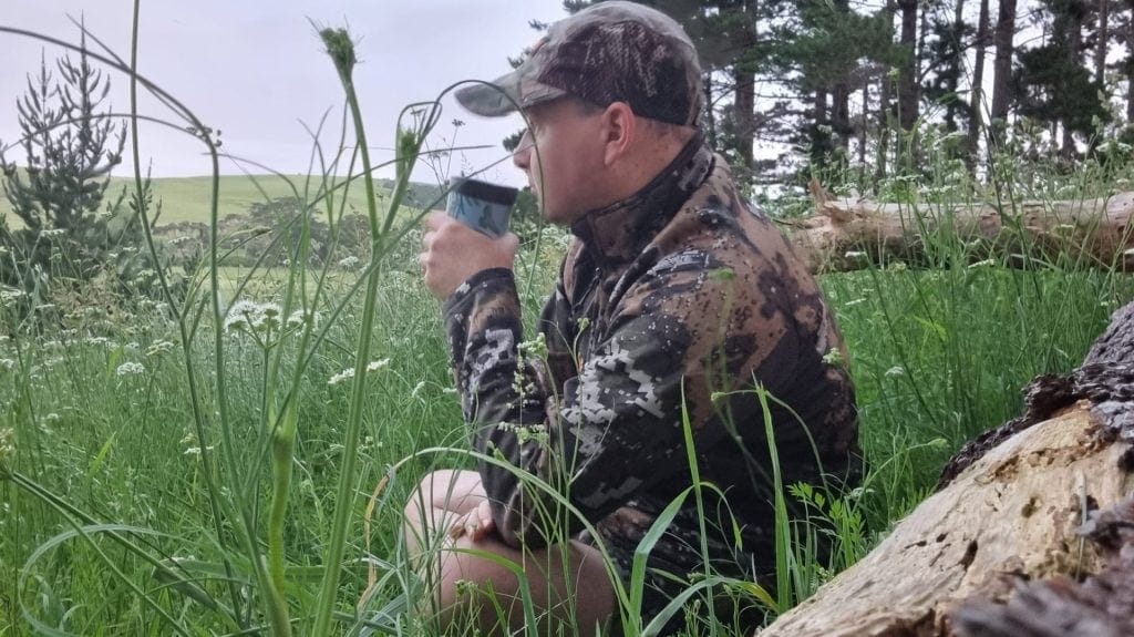 Person wearing camo clothes sitting in high grass with trees in background